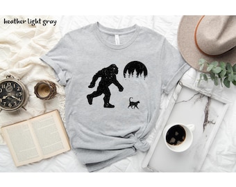 May include: Heather light gray t-shirt featuring a black silhouette of Bigfoot walking, a black cat, and a forest scene under a full moon. The shirt is a crew neck style.