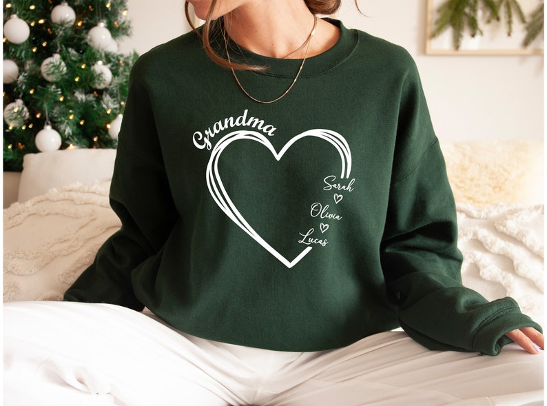 Pu&ograve; includere: Felpa verde con un design a forma di cuore bianco e il testo "Grandma" sopra il cuore. All'interno del cuore ci sono i nomi "Sarah", "Olivia" e "Lucas" con piccoli cuori accanto a ciascun nome.