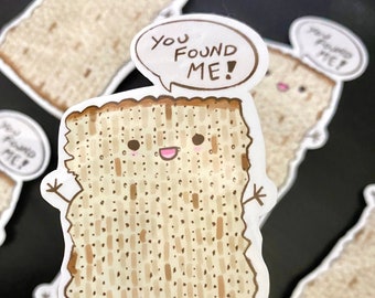 May include: A set of four matzah stickers with a cartoon matzah character with a speech bubble that says "You found me!" The matzah is brown and beige with a grid pattern.
