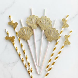 May include: A collection of decorative straws with gold glitter seashell and bone-shaped toppers. The straws have white and gold stripes. Some straws have a pearlescent finish. The items are arranged on a white marble surface.