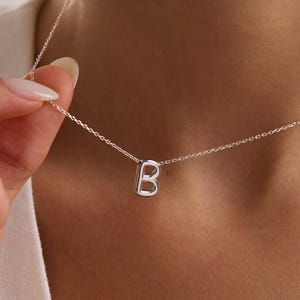 May include: A silver necklace with a small, capital letter "B" pendant. The pendant is suspended from a fine chain, offering a minimalist and elegant aesthetic. The necklace is displayed against a neutral backdrop.