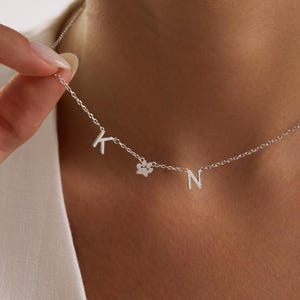 Puede incluir: Un collar plateado con las letras "K" y "N" y un dije de huella de pata. La delicada cadena es sostenida por una persona. El collar es una pieza de joyería personalizada.
