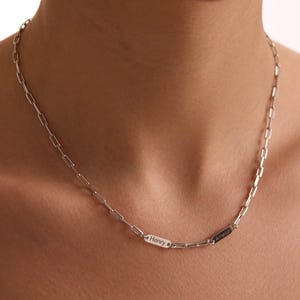 Può includere: Una collana a catena color argento con due piccoli ciondoli rettangolari. Uno recita "Henry" e l'altro "Amelia". La collana è indossata intorno al collo.