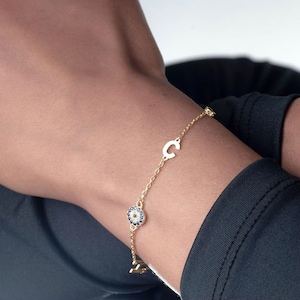May include: A gold chain bracelet with a small, round charm featuring a blue and white design, and a gold letter "C" charm. The bracelet is worn on a person's wrist, against a dark blue sleeve.