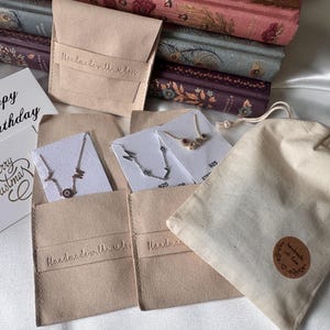 Op de afbeelding: Een assortiment sieraden, waaronder kettingen, gepresenteerd in kleine beige zakjes en cadeaukaarten. De zakjes zijn voorzien van het opschrift "handmade with love". De kaarten luiden "Happy Birthday" en "Merry Christmas". Een kleine canvas tas met een ronde sticker is ook zichtbaar.