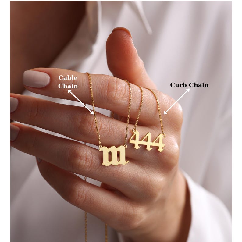 May include: Two gold necklaces with different chain styles. The necklace on the left is a cable chain with a pendant that says "M" in old English font. The necklace on the right is a curb chain with a pendant that says "444" in old English font.