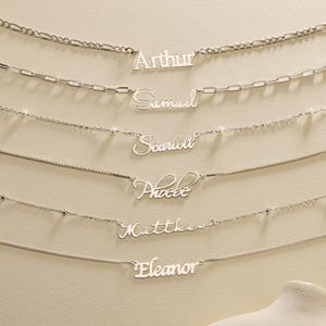 Könnte beinhalten: Silberne Namensketten mit verschiedenen Kettenstilen. Die Namen "Arthur", "Samuel", "Scarlett", "Phoebe", "Matthew" und "Eleanor" sind in Schreibschrift dargestellt. Die Halsketten werden auf einem neutralen Hintergrund präsentiert.