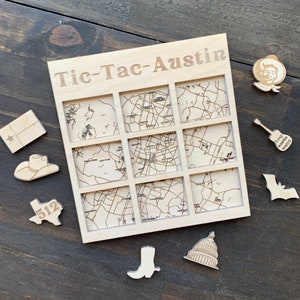 May include: A wooden Tic-Tac-Toe board with the text "Tic-Tac-Austin" engraved on it. The board features nine squares with engraved maps of Austin, Texas. Surrounding the board are wooden cutouts of Texas symbols, including a cowboy hat, the state flag, and the state capitol.