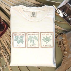 May include: Cream-colored t-shirt with three postage stamp designs featuring Hawaiian plants and the words "Hawaii" above each. The stamps display "'ULU", "KALO", and "LAU KI" in a light green color. The shirt is a size XL.