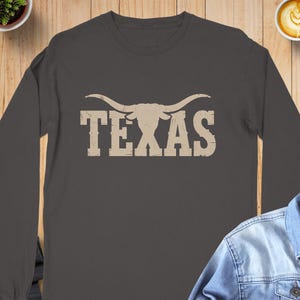 May include: Dark gray long-sleeve shirt featuring the word "TEXAS" in a distressed, tan font, with a longhorn silhouette above the text. The shirt is made of a soft material.