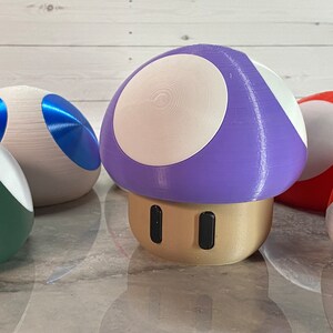 May include: A collection of 3D printed mushroom-shaped objects. The mushrooms are in various colors, including purple, blue, green, and red. The mushrooms have white tops and are sitting on a light gray surface.