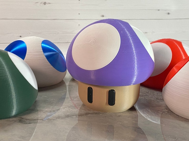 May include: A collection of 3D printed mushroom-shaped objects. The mushrooms are in various colors, including purple, blue, green, and red. The mushrooms have white tops and are sitting on a light gray surface.