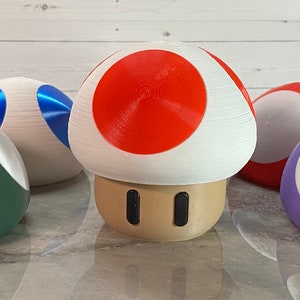 May include: A set of five 3D printed mushroom-shaped figurines. The mushrooms are in various colors, including red, white, blue, green, and purple. The mushrooms are all about 3 inches tall.