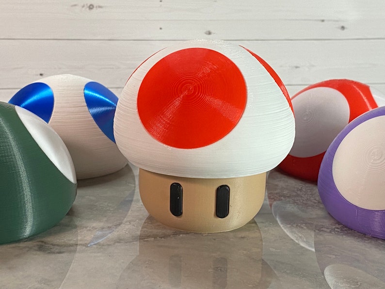 May include: A set of five 3D printed mushroom-shaped figurines. The mushrooms are in various colors, including red, white, blue, green, and purple. The mushrooms are all about 3 inches tall.