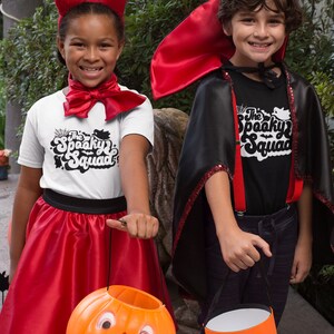 May include: Two children dressed in Halloween costumes. One child is wearing a red devil costume with horns and a red skirt. The other child is wearing a black vampire costume with a red cape. Both children are holding orange pumpkin-shaped trick-or-treat buckets. The children are smiling and looking at the camera. The text on their shirts says "The Spooky Squad".