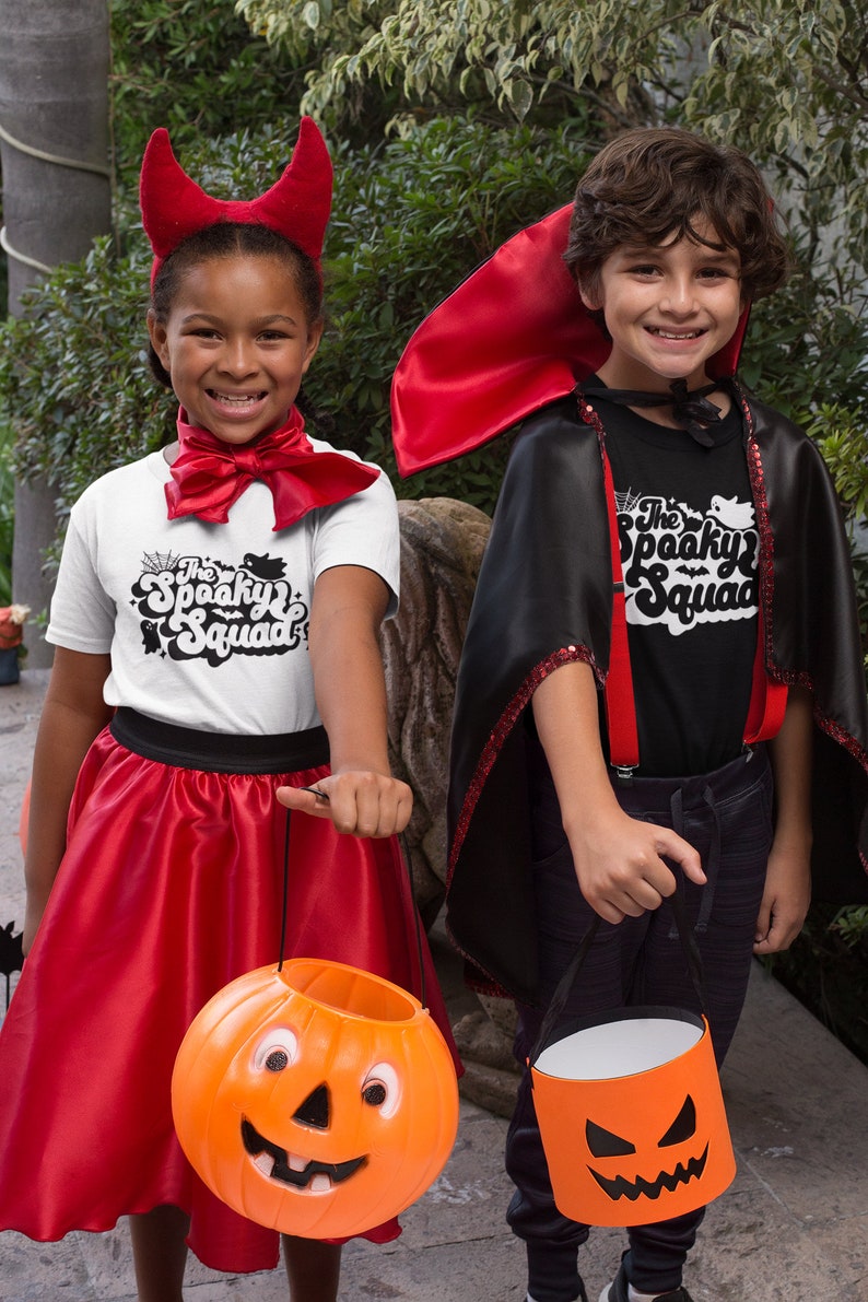 May include: Two children dressed in Halloween costumes. One child is wearing a red devil costume with horns and a red skirt. The other child is wearing a black vampire costume with a red cape. Both children are holding orange pumpkin-shaped trick-or-treat buckets. The children are smiling and looking at the camera. The text on their shirts says "The Spooky Squad".