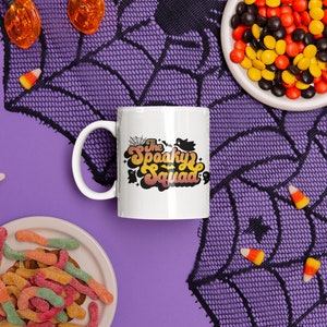 May include: White ceramic coffee mug with a black and orange design that says "The Spooky Squad". The mug is on a purple background with a black spiderweb and candy corn.