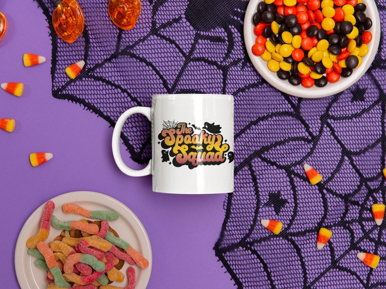 May include: White ceramic coffee mug with a black and orange design that says "The Spooky Squad". The mug is on a purple background with a black spiderweb and candy corn.