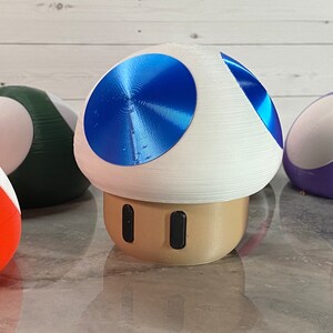 May include: A collection of 3D printed mushroom figurines. The mushrooms are various colors, including red, green, purple, and blue. The blue mushroom has a shiny, metallic finish.