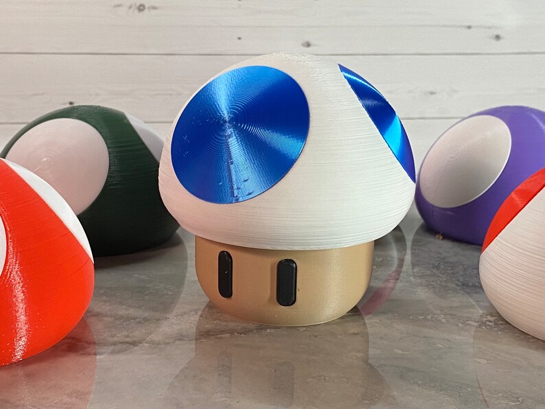 May include: A collection of 3D printed mushroom figurines. The mushrooms are various colors, including red, green, purple, and blue. The blue mushroom has a shiny, metallic finish.