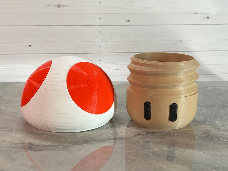 May include: A 3D printed mushroom-shaped object with a red top and white bottom, and a 3D printed brown cylinder with two black circles.