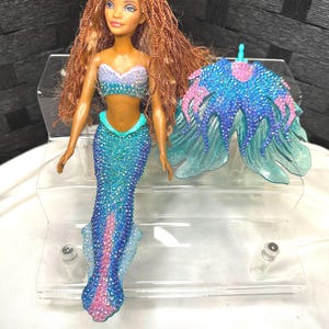May include: A mermaid doll with long, braided auburn hair, wearing a blue and purple sequined top and a matching sequined tail. A separate, blue and pink sequined accessory is also included. The doll is displayed on a clear, plastic stand.