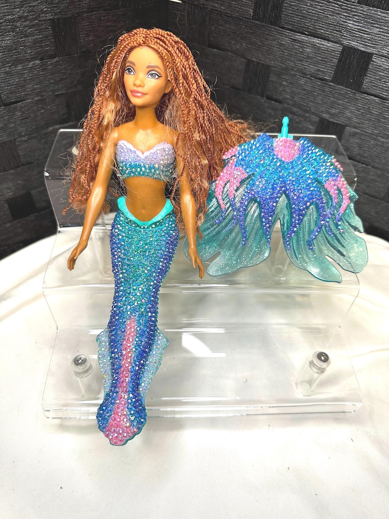 May include: A mermaid doll with long, braided auburn hair, wearing a blue and purple sequined top and a matching sequined tail. A separate, blue and pink sequined accessory is also included. The doll is displayed on a clear, plastic stand.