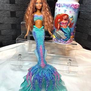 May include: A mermaid doll with auburn hair, wearing a turquoise and purple top and a matching mermaid tail. A tumbler with a mermaid graphic and blue jeweled accents is next to the doll.