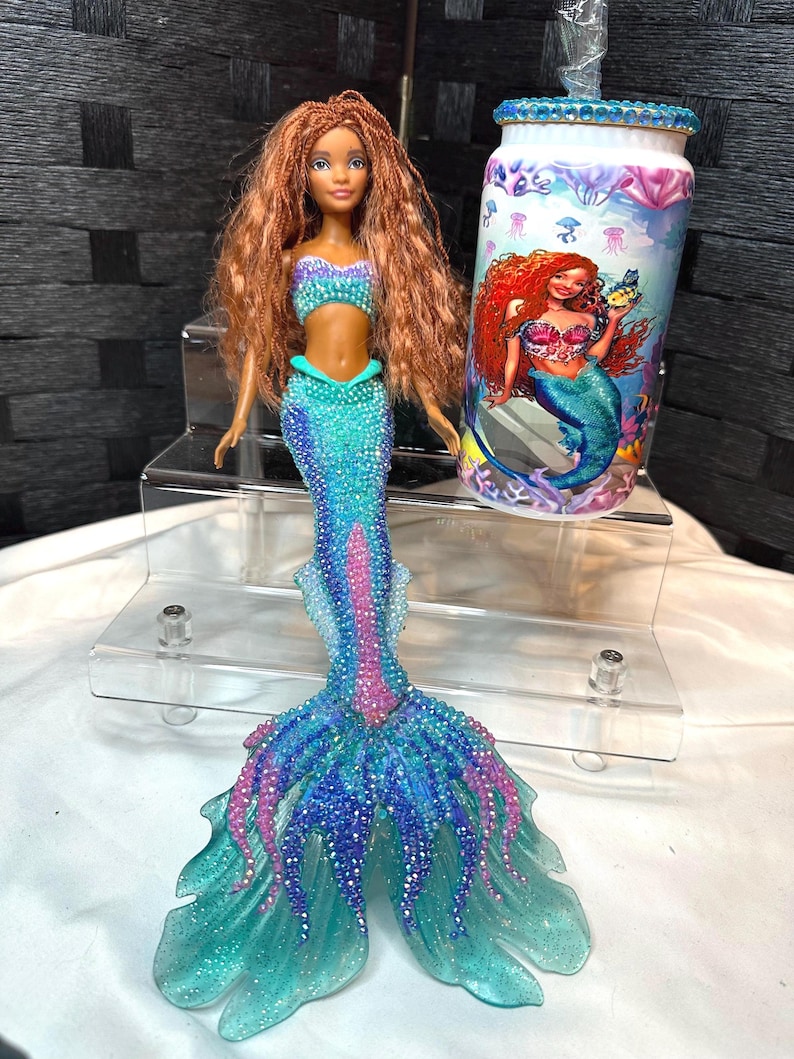 May include: A mermaid doll with auburn hair, wearing a turquoise and purple top and a matching mermaid tail. A tumbler with a mermaid graphic and blue jeweled accents is next to the doll.