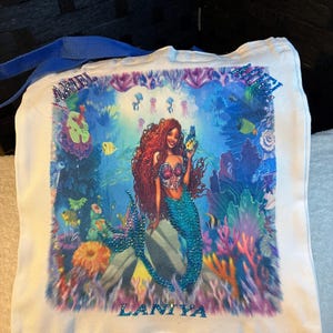 May include: White canvas tote bag with a mermaid illustration. The mermaid has red hair and a turquoise tail, surrounded by colourful coral and fish. The name "LANIYA" is displayed in blue rhinestones.