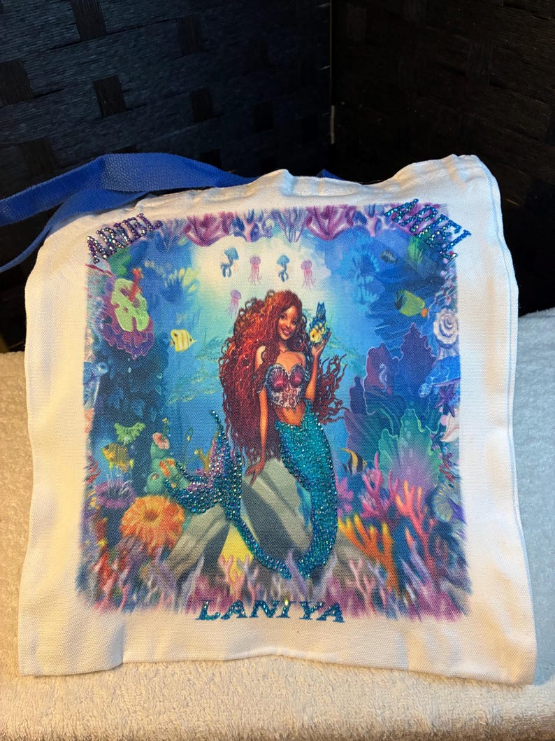 May include: White canvas tote bag with a mermaid illustration. The mermaid has red hair and a turquoise tail, surrounded by colourful coral and fish. The name "LANIYA" is displayed in blue rhinestones.