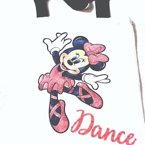 Puede incluir: Bolso tote blanco con una Minnie Mouse de dibujos animados con un atuendo de ballet rosa. La palabra "Dance" está escrita en rosa debajo del personaje. Un diseño "NY" negro en la parte superior.