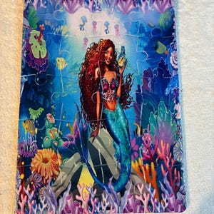 May include: A colourful jigsaw puzzle depicting a mermaid in an underwater scene. The mermaid has long red hair, a blue tail, and holds a glowing object. The puzzle includes fish, coral, and other sea creatures. The puzzle is rectangular.