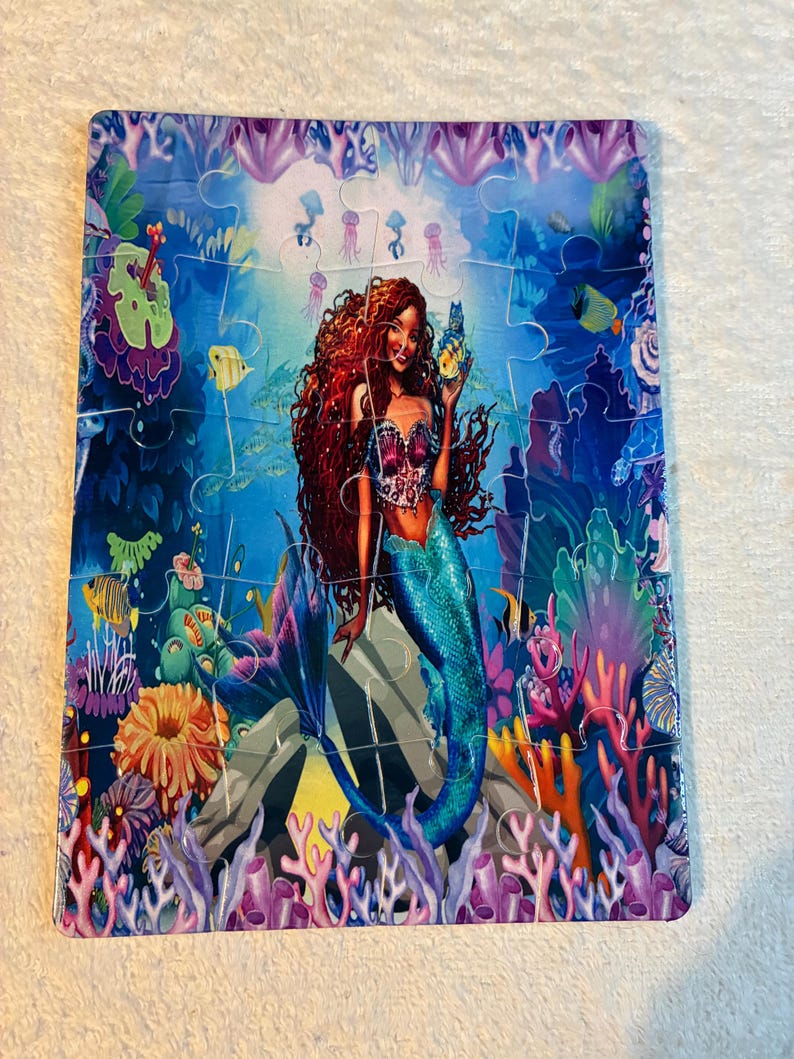 May include: A colourful jigsaw puzzle depicting a mermaid in an underwater scene. The mermaid has long red hair, a blue tail, and holds a glowing object. The puzzle includes fish, coral, and other sea creatures. The puzzle is rectangular.