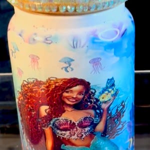 May include: A cylindrical tumbler with a mermaid design. The tumbler has a light blue and yellow background with illustrations of jellyfish and sea plants. The mermaid has red hair and a blue tail. The lid is decorated with iridescent rhinestones.