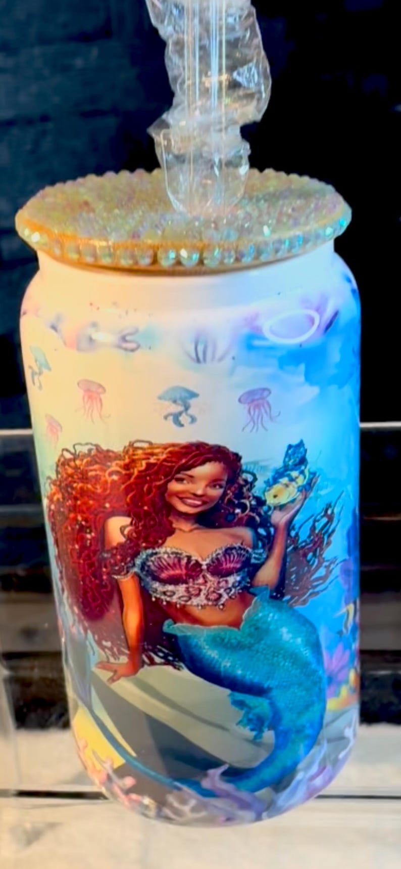 May include: A cylindrical tumbler with a mermaid design. The tumbler has a light blue and yellow background with illustrations of jellyfish and sea plants. The mermaid has red hair and a blue tail. The lid is decorated with iridescent rhinestones.