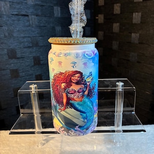 May include: A cylindrical tumbler with a mermaid design. The mermaid has red hair and a blue tail, holding a small fish. The tumbler has a clear straw and a decorative lid with rhinestones. The background has a blue and purple watercolor effect.