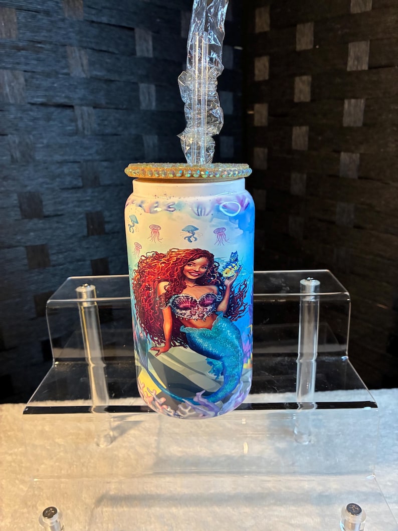 May include: A cylindrical tumbler with a mermaid design. The mermaid has red hair and a blue tail, holding a small fish. The tumbler has a clear straw and a decorative lid with rhinestones. The background has a blue and purple watercolor effect.