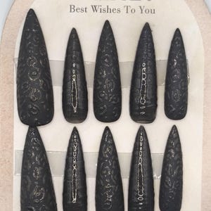 May include: Set of ten black, stiletto-shaped false nails with a textured, embossed design. The nails are arranged on a cardboard backing with the text "Nail Art Best Wishes To You".