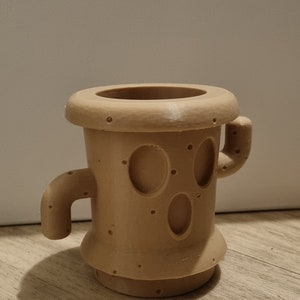 May include: A tan-coloured, cylindrical ceramic mug with a unique design. It features a wide rim, two small handles, and a series of small holes and oval cutouts resembling a face. The mug is set on a wooden surface.
