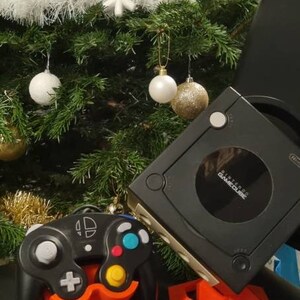 Könnte beinhalten: Eine schwarze Nintendo GameCube Konsole und ein passender schwarzer Controller, die jeweils auf einem individuellen orangefarbenen Kunststoffständer stehen. Die Konsole hat eine runde Disc-Abdeckung mit dem Nintendo GameCube Logo.