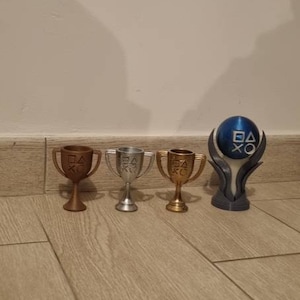 Puede incluir: Una colección de cuatro objetos con forma de trofeo. Tres son metálicos, en bronce, plata y oro. El cuarto es una esfera azul y blanca con un soporte. Todos están sobre una superficie de madera.
