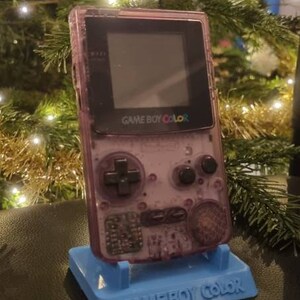 Könnte beinhalten: Eine durchscheinende violette Game Boy Color Handheld-Spielkonsole mit schwarzem Bildschirmrahmen und schwarzen Tasten, die auf einem hellblauen Kunststoff-Präsentationsständer mit dem geprägten Text GAME BOY COLOR steht.