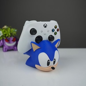 May include: A blue Sonic the Hedgehog figure holding a white Xbox controller. The controller has black buttons and colored action buttons. The Sonic figure has a tan muzzle and white eyes. A purple Pokemon figure is in the background.
