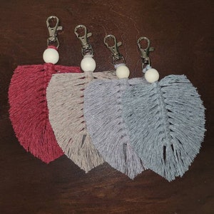 Peut inclure: Quatre porte-clés en macramé en forme de plumes, dans les tons de rouge, beige, gris et bleu. Chaque porte-clés est doté d'un fermoir argenté et d'une perle en bois blanche. Les plumes sont nouées de manière complexe, créant un aspect texturé.