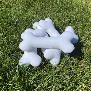 May include: A pile of white, plush dog bone toys resting on a bed of green grass. The bones are soft and fluffy, with a classic bone shape. The toys are designed for pets.