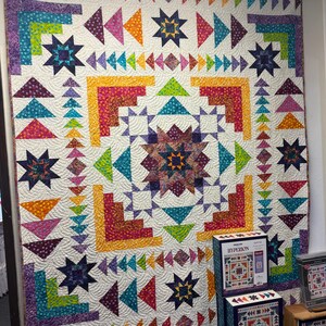 May include: A large, square quilt featuring a central star motif surrounded by geometric log cabin blocks and triangular patterns in vibrant shades of purple, teal, yellow, orange, and pink on a white background, with Hyperion quilt kit boxes.