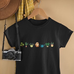 Może przedstawiać: Czarny T-shirt z grafiką doniczkowych roślin i małego psa. Pies siedzi w żółtej doniczce i trzyma konewkę.