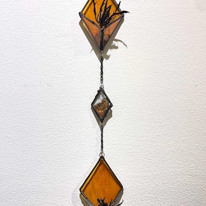 May include: A hanging stained glass art piece featuring three diamond-shaped panels. The top and bottom panels are amber-colored with black botanical elements inside. The middle panel is clear with gold flecks. All panels are connected by a chain.