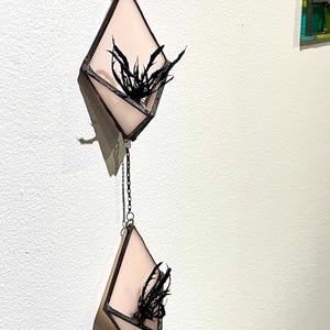 May include: Two hanging geometric stained glass planters. Each diamond-shaped planter features a pale pink glass panel and black metal framing. Black, feathery accents are inside the planters. The planters are connected by a silver chain.